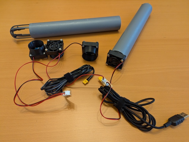 Four 40 mm fans with the holder and a fan guard attached scattered on the desk, one with a 40 mm tube attached, another tube and a fitting leaf guard as well as assorted cables and connectors lying around.
