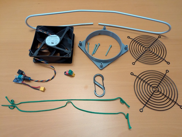 Assorted components on the desk: a aluminum loop, a fan, a fan mount, screws, two fan guards, a PWM controller, a USB C trigger module, two short cords, a double carabiner.