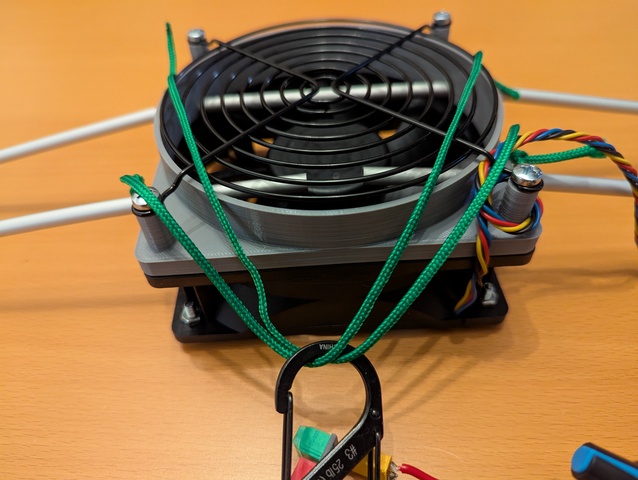 Detail of the mount with the funnel and fan guard clamping the aluminum loop on the fan, two green cords are attached to the loop, a carabiner hook attached to the cords.