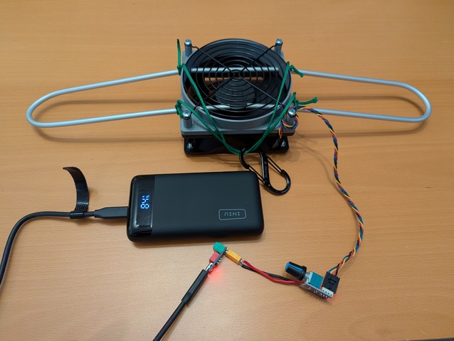 On a desk lies an aluminum rod bent to a loop with a grey 3D printed mount on top clamping a 120 mm fan below. The mount has a nozzle with a fan guard on top. The fan is connected to a PWM controller adjustable by a potentiometer and powered by a USB C power bank with the 'trigger module' setting it to 12V.